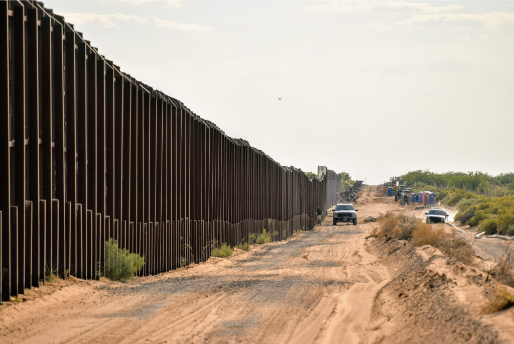 long view of the border wall between the usa and mexico with a couple trucks driving along side it